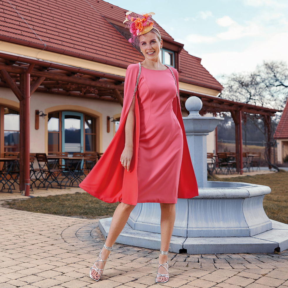 Mode: Frau in himbeereis-färbigen Etuikleid mit passendem Umhang mit Strassborte und Fascinator-Hut mit orangen und pinken Blumen und Schleier.