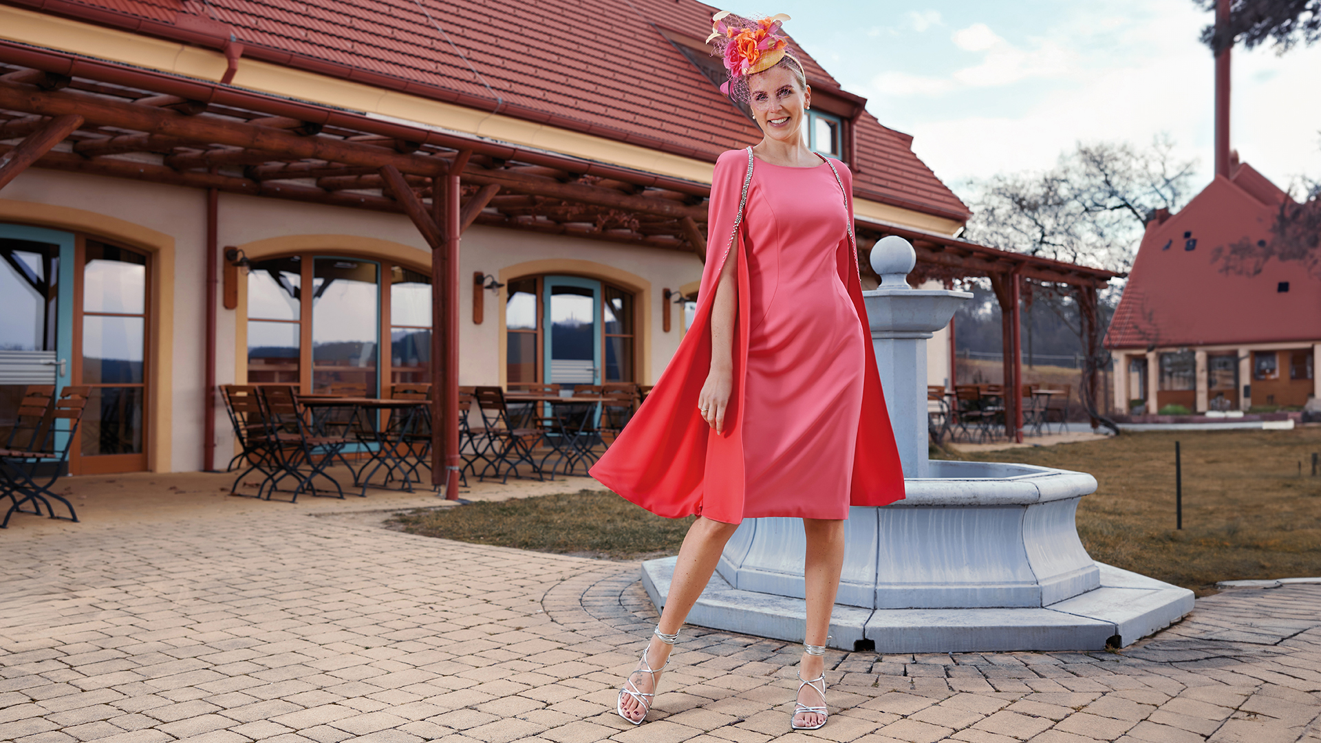 Mode: Frau in himbeereis-färbigen Etuikleid mit passendem Umhang mit Strassborte und Fascinator-Hut mit orangen und pinken Blumen und Schleier.