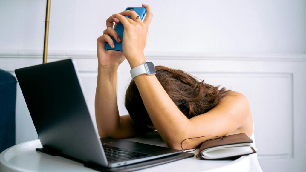 Frau hat beide Ellenbogen auf einen Tisch gestützt, ihr Kopf liegt nach unten gerichtet auf dem Tisch. Vor ihr steht ein Laptop. in ihren Händen hält sie ein Handy.