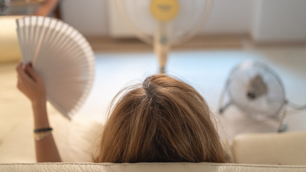 Hitzewallungen: Frau mit sitzt auf einer Couch mit einem Fächer in der Hand und 2 Ventilatoren vor sich