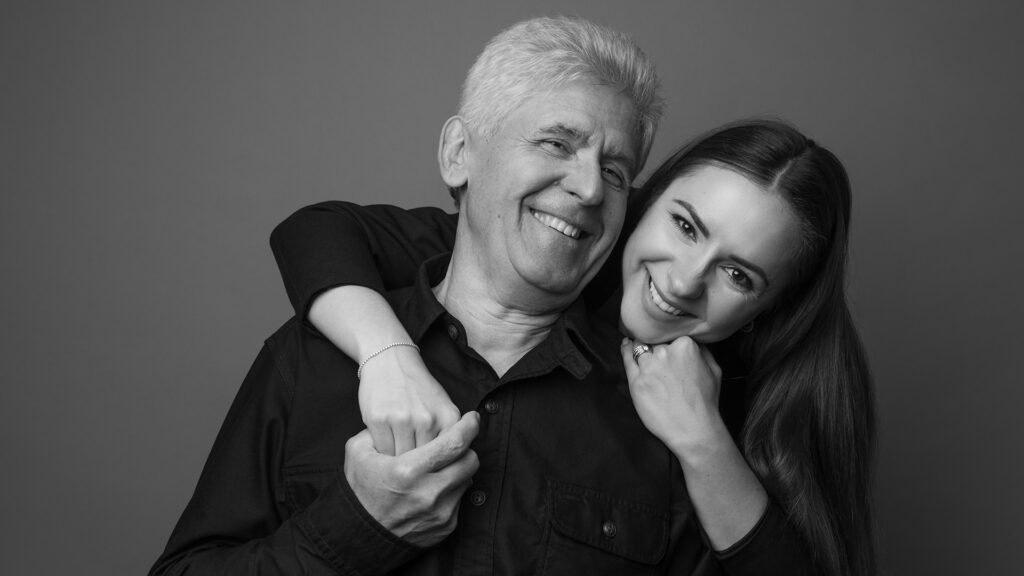 Studio portrait of a Ukrianian father in his 60s and his adult daughter in her late 20s. They are both wearing black and the daughter is hugging her father. The photo is in black and white.