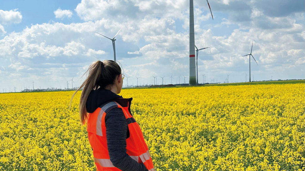 Studentin: Junge Frau mit neon-oranger Warnweste steht vor einem Rapsfeld mit 4 Windrädern im Hintergrund
