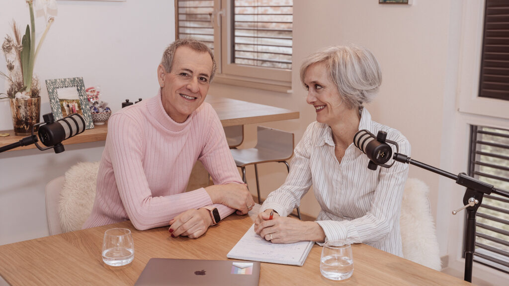 Transition: Ein geschminkter Mann mit roten Fingernägeln schaut in die Kamera und die Frau die mit ihm am Tisch sitzt lächelt ihn an. Im Vordergrund zwei Microfone.