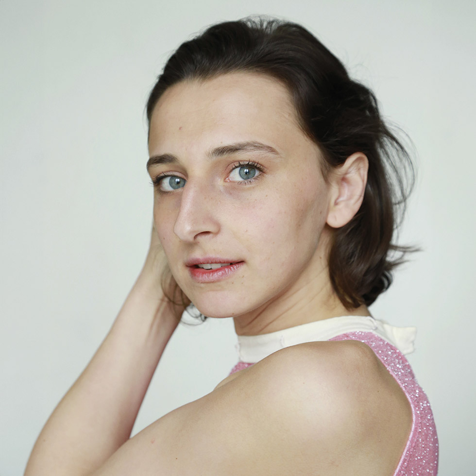 Clara Wolfram: Portrait einer jungen Frau mit großen blau-grauen Augen die über die Schulter in die Kamera blickt