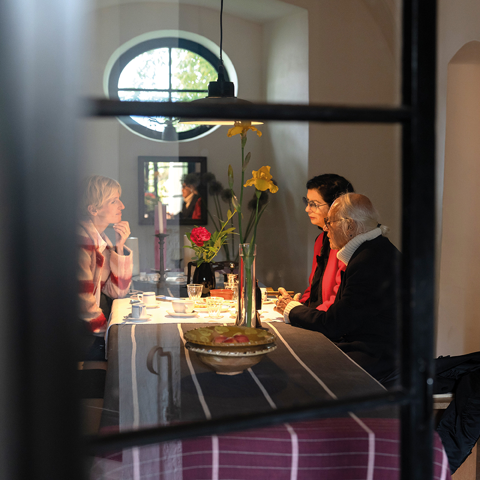 3 Menschen an einem gedeckten Tisch unterhalten sich – durch's Fenster fotografiert