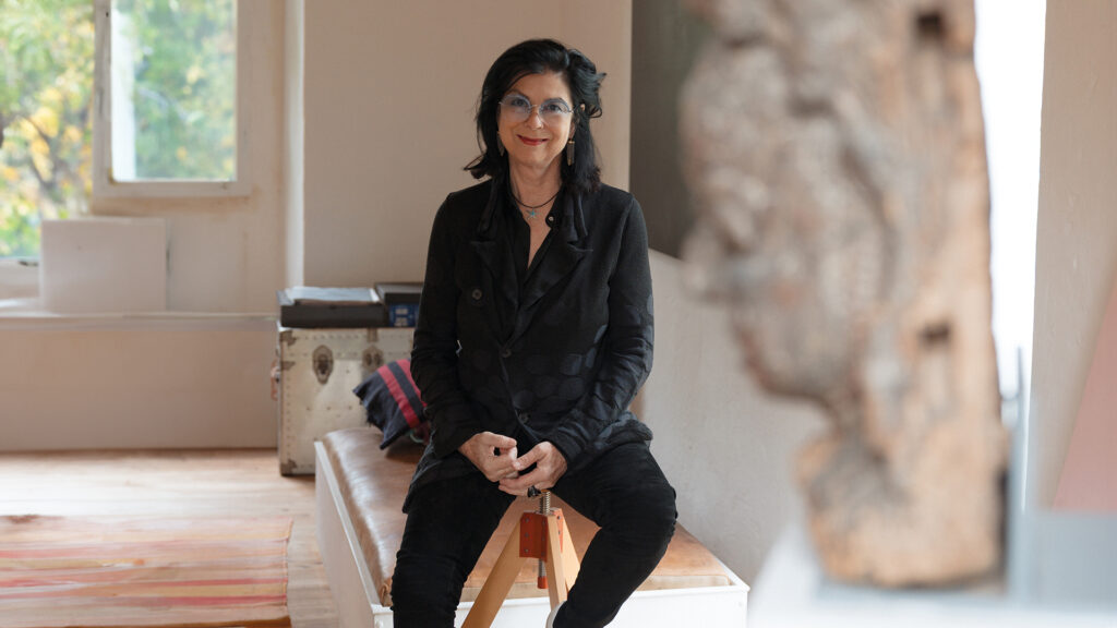 Portrait von Eva Schlegel – schwarzhaarige lächelnde Frau im mittleren Alter und blau getönter Brille sitzt auf einem Sessel ohne Lehne