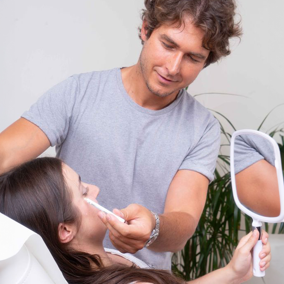 OmniMed: Mann zeigt einer in einem Behandlungssessel liegenden Frau, die einen Handspiegel hält, mit einem Stift zum Auge