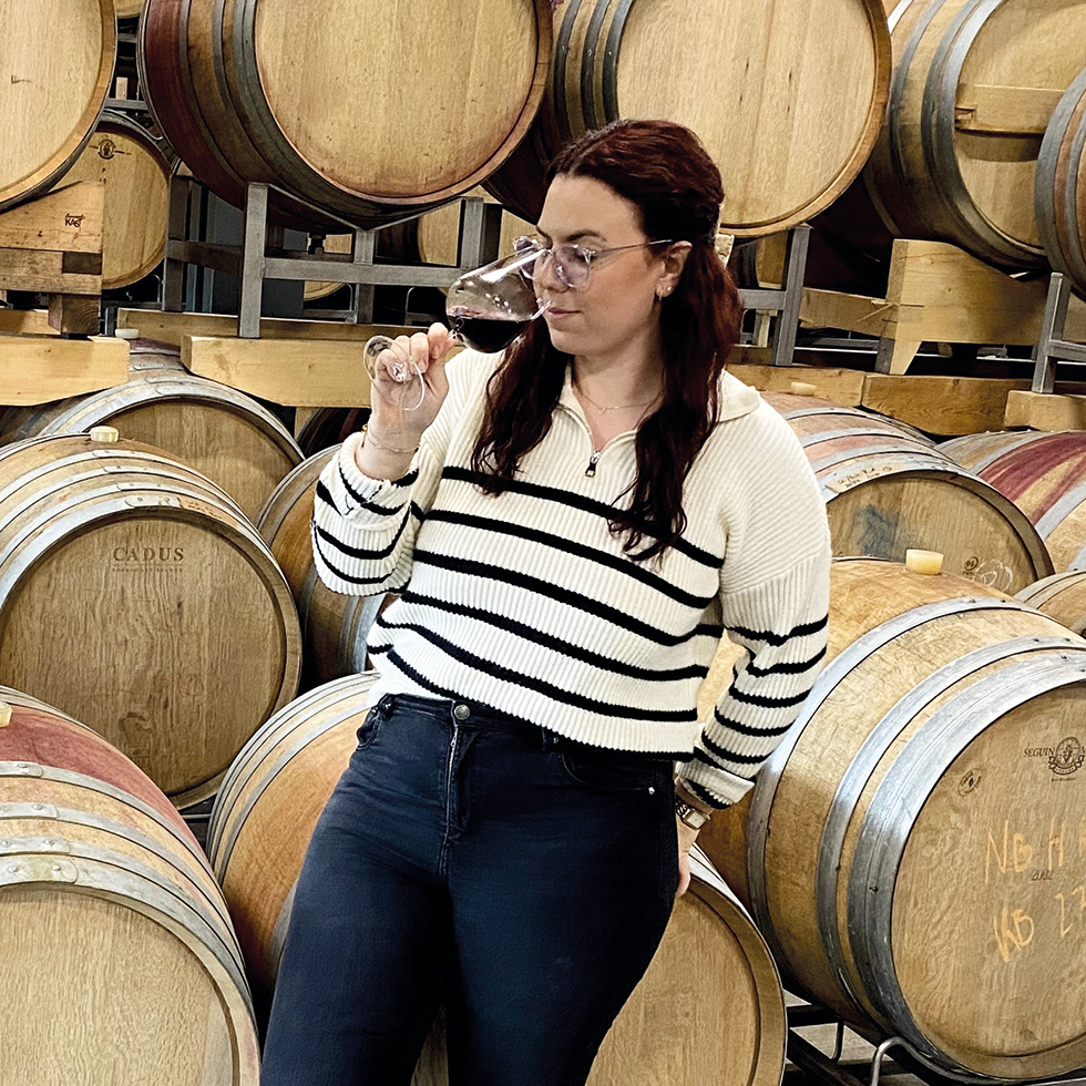 Junge Dame verkostet einen Rotwein, im Hintergrund Weinfässer aus Holz