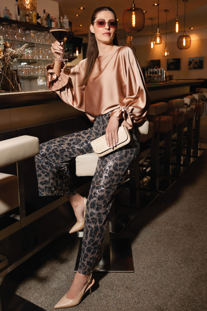 Sommermode: Frau in Seidenbluse und Leopardenprint-Hose an einer Bar mit Cocktail in der Hand