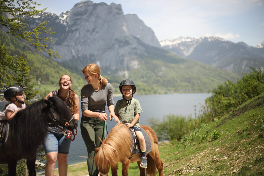 Narzissendorf Zloam: 2 Kinder mit Helm auf Ponys und dabei 2 lachende junge Damen