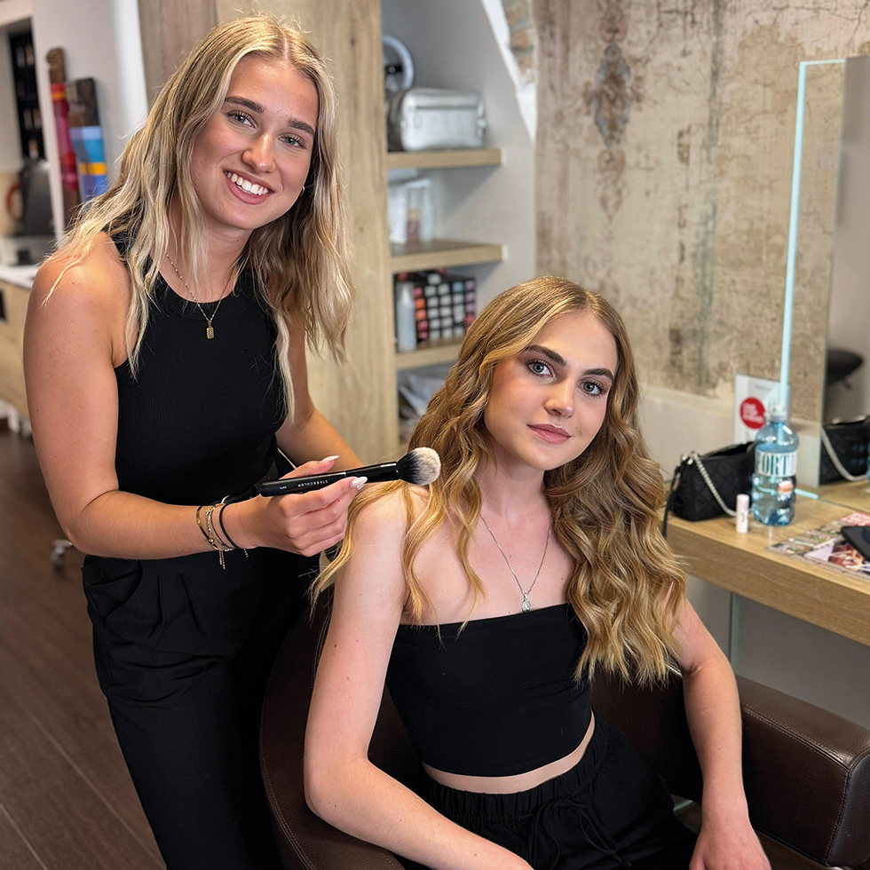 Eine Blonde Make-up-Artistin schminkt eine dunkelblonde junge Frau, die vor ihr am Stuhl sitzt.