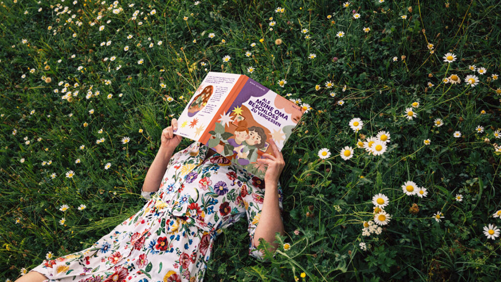 Frau mit sommerlichen Blümchenkleid liegt in der Wiese mit einem Buch vor dem Gesicht