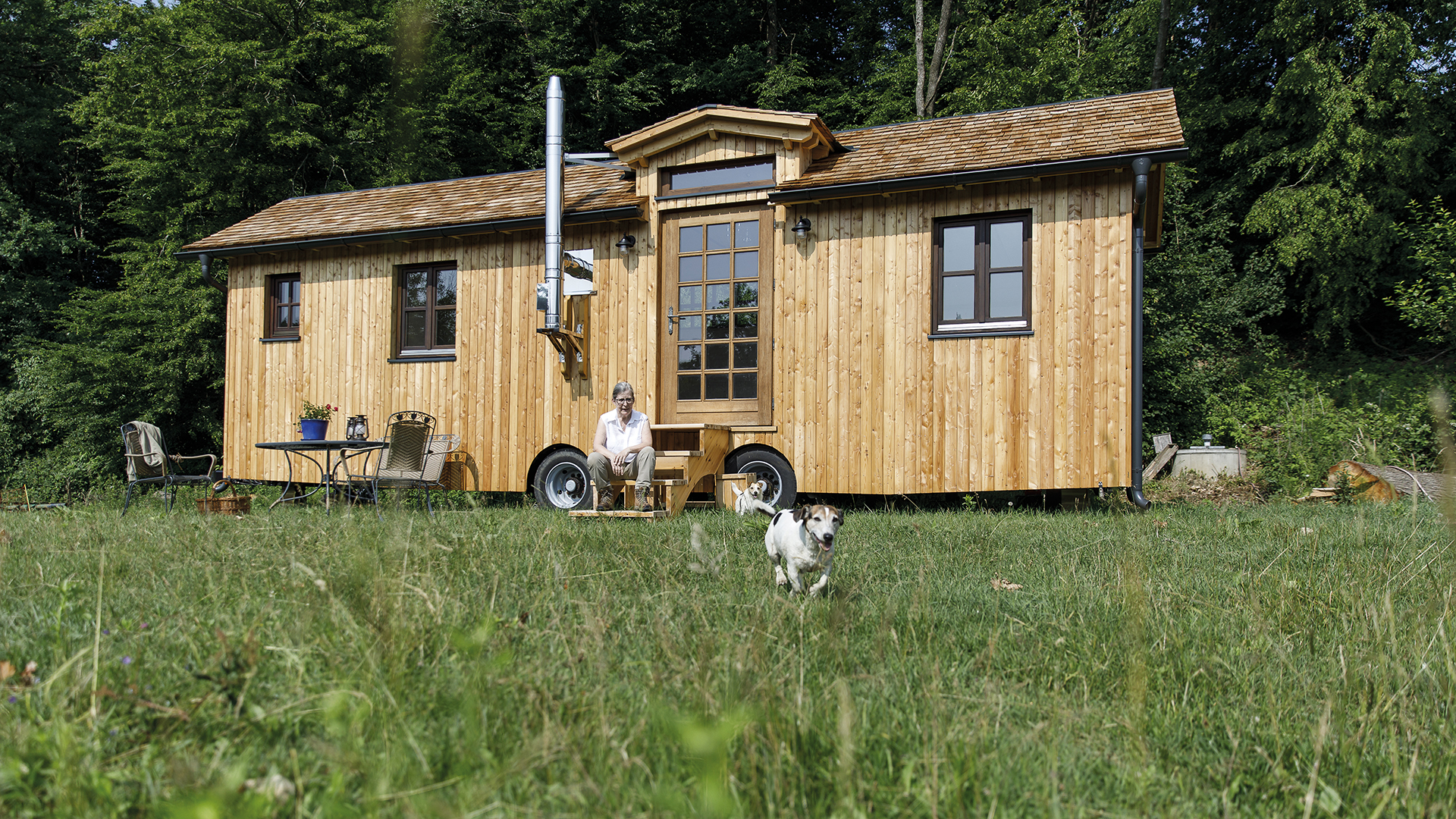 Judith Anger: Mobile Home aus Holz – auf den Treppen sitzt eine Frau in Vordergrund läuft ein kleiner Hund auf einen zu.