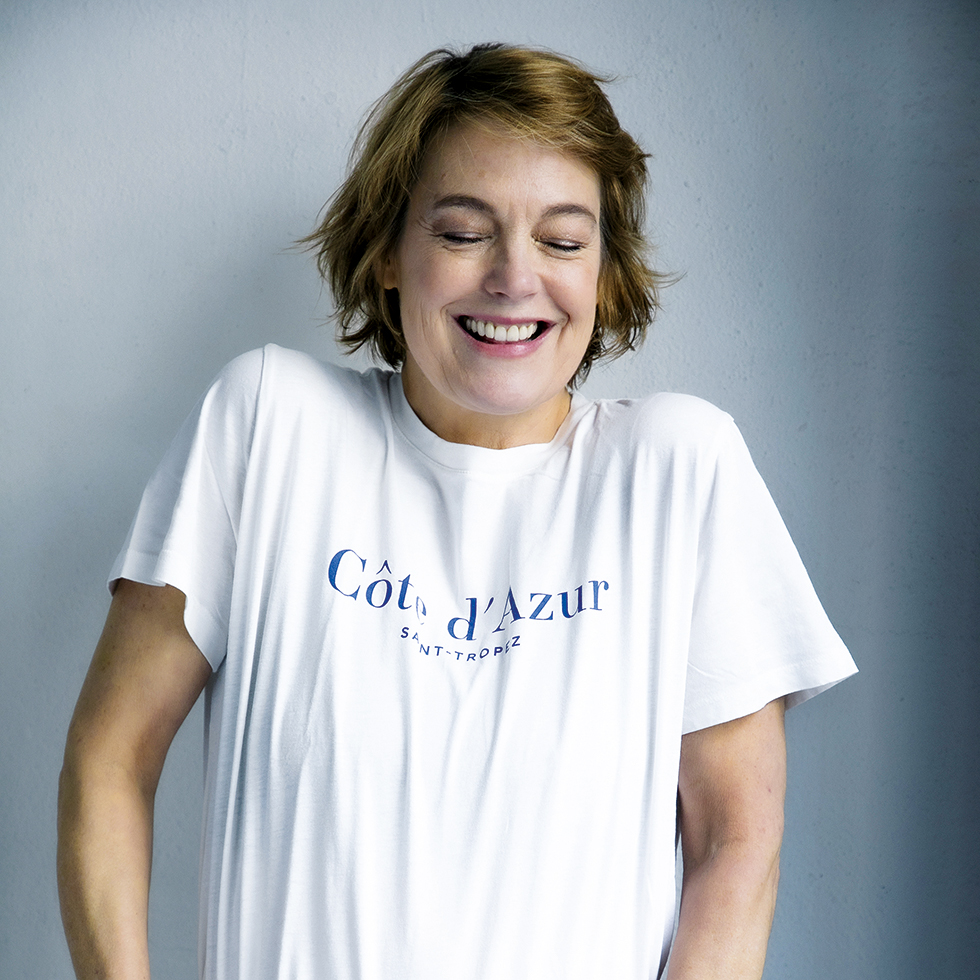 Elke Winkens: Frau in weißem T-Shirt mit der Aufschrift Cote d'Azur Saint-Tropez zieht lachend mit geschlossenen Augen die Schultern hoch