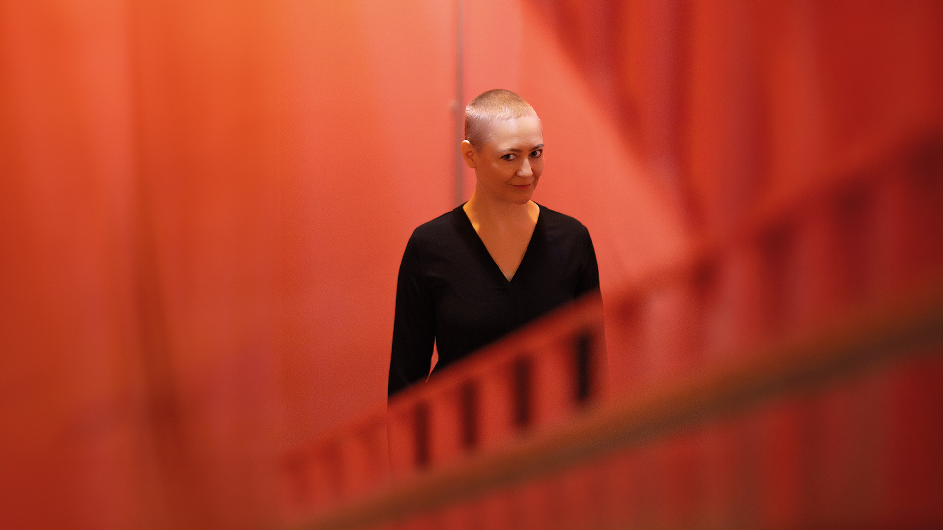 Petra Piuk: Frau mit kurzgeschorenem Haar vor Roter Wand schaut einen schräg von der Seite an