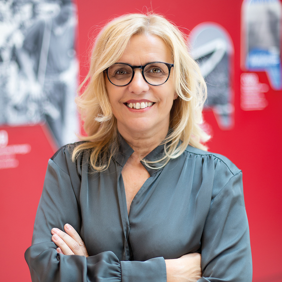 Stadtvilla: Portrait einer blonden älteren Frau mit schwarzer Brille und verschränkten Armen
