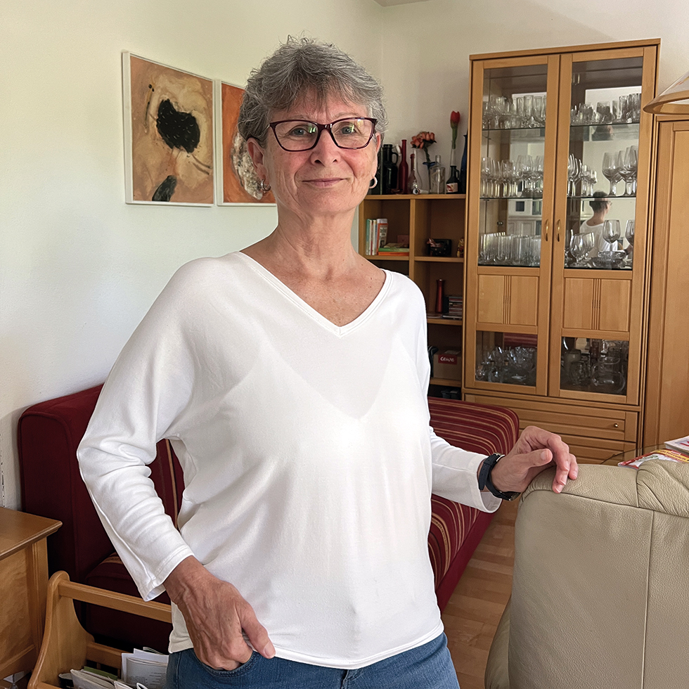 Verein HPE: Ältere Dame in weißem, langärmeligen T-Shirt steht vor einem Wohnzimmerschrank