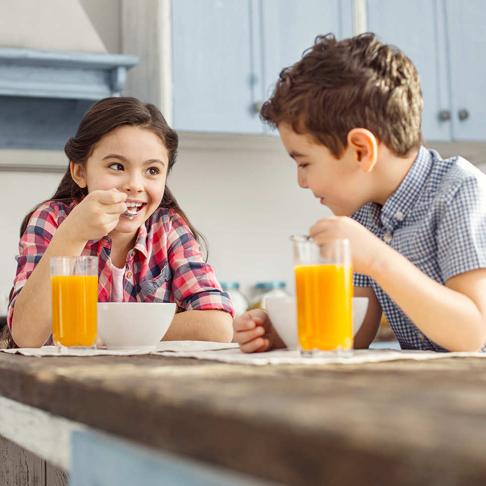 Hübsches, inspiriertes kleines dunkelhaariges Mädchen lächelt und schaut ihren Bruder an, während sie frühstücken.“