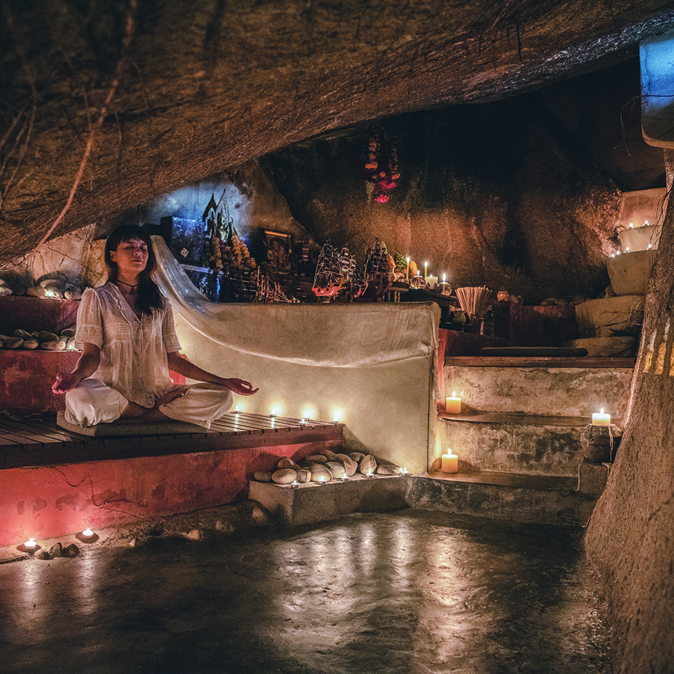 Höhle mit vielen Kerzen und einer Frau im weißen Kleid im Lotus-Sitz meditierend