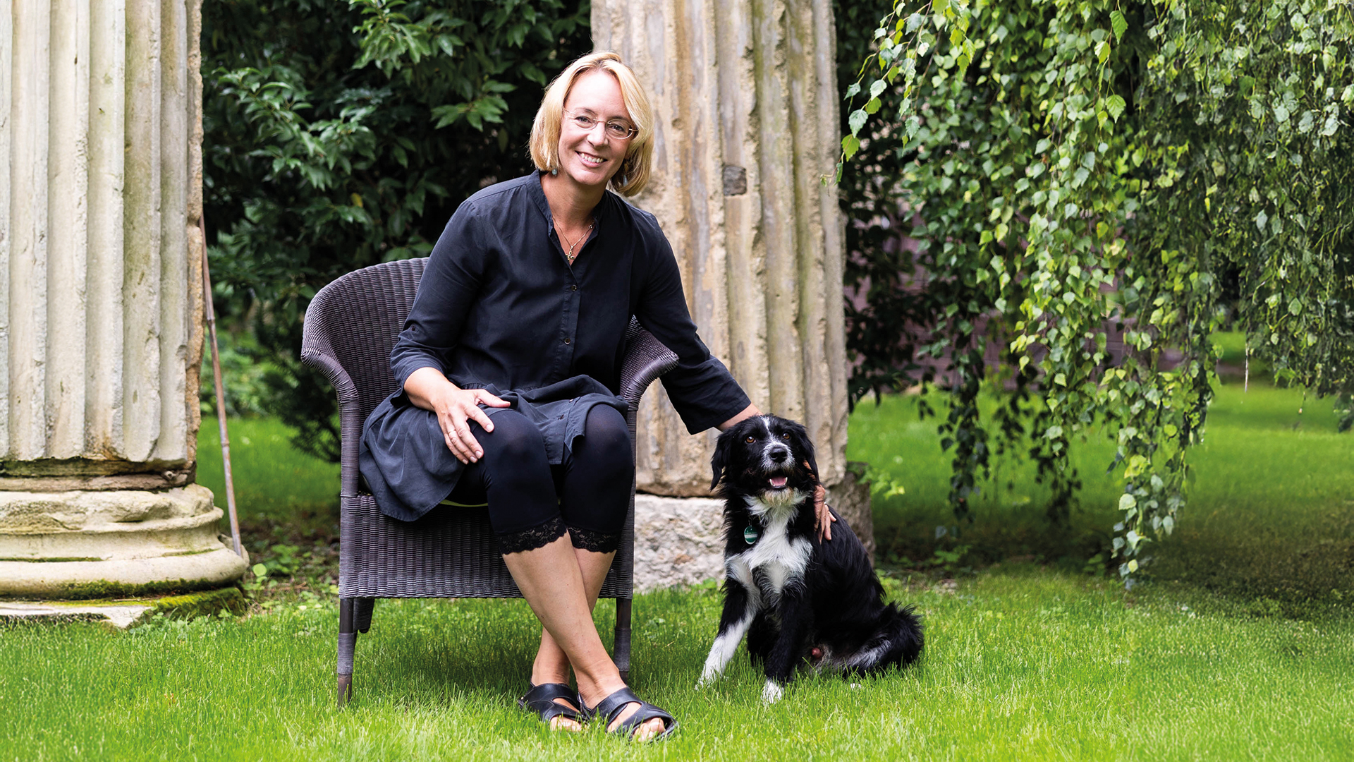 Sarah Spiekermann: Frau in dunkelblauem Kleid und Brille sitzt auf einem Sessel im Garten und streichelt einen Bordercollie-Mix