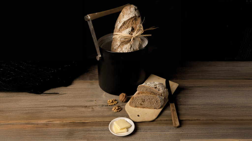 Vollkorn-Nussbrot: Ein länglicher Laib Brot in einem schwarzen Blechkübel davor ein Holzbrett mit 2 aufgeschnittenen Scheiben Walnussbrot