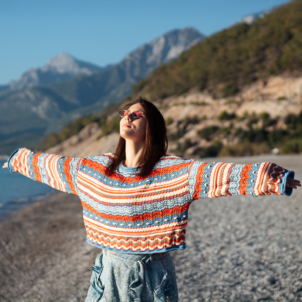 Frau in einem hellen Strickpullover und rosa Sonnenbrille genießt die Sonne am Strand mit Bergen im Hintergrund.