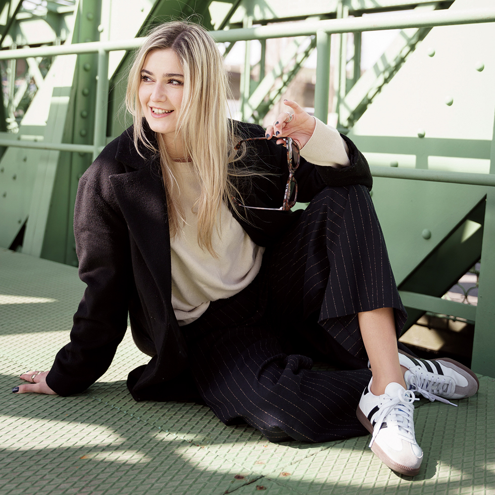 Vorfreude: Blonde junge Dame im Hosenanzug uns Sneakers sitzt auf einer Stahbrücke