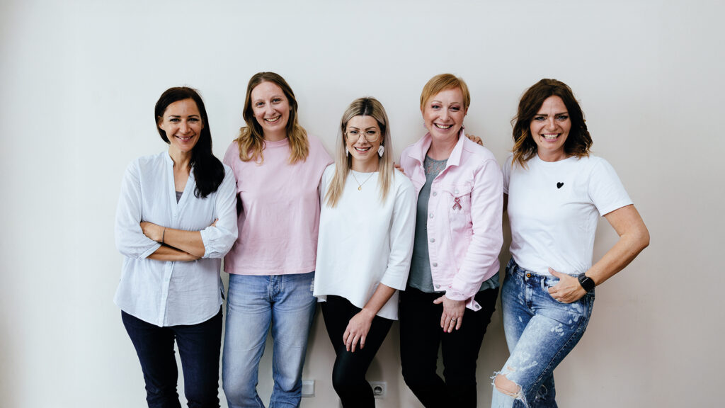 Feel again: 5 Frauen mit Pink-Ribbon-Schleife stehen vor einer Wand und lächeln