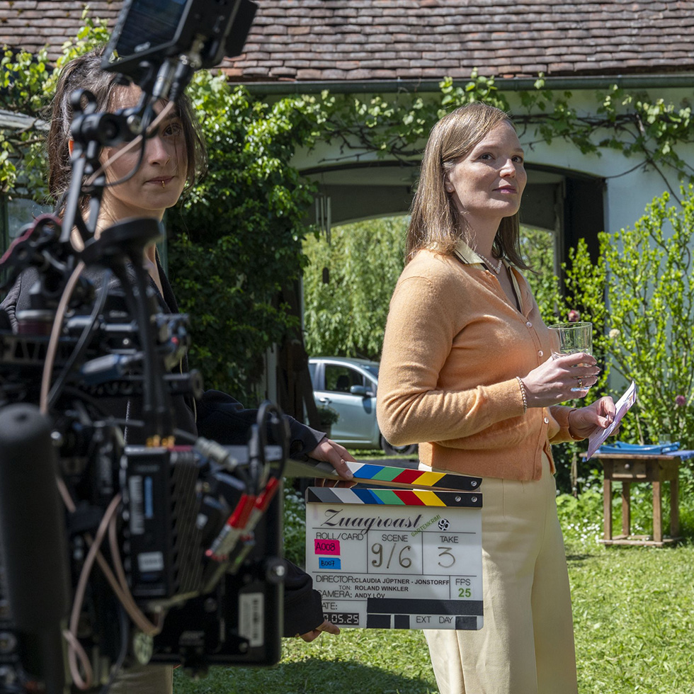 Zuagroast: Filmszene im Garten mit Klappe – Eine Frau im orangefarbenen Cardigan steht mit einem Glas Wasser, daneben hält jemand eine Filmklappe; im Vordergrund Kameraausrüstung.