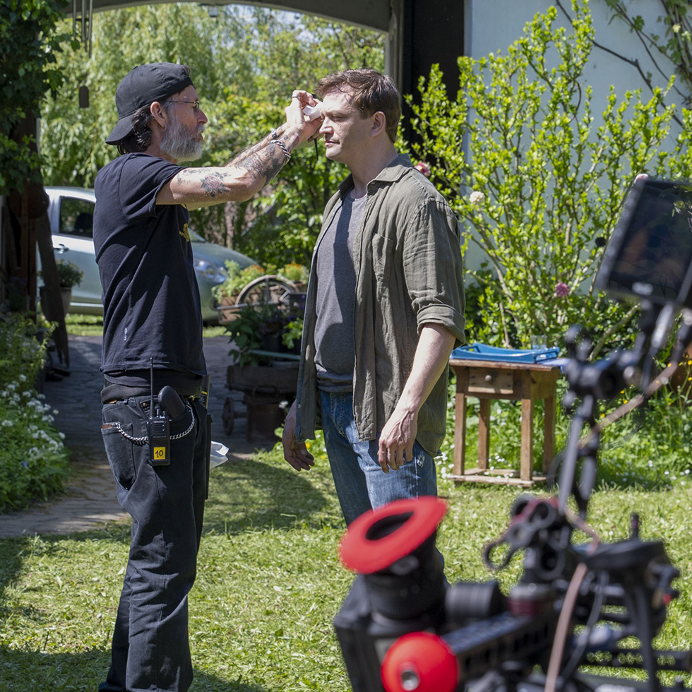 Make-up am Set – Ein Crew-Mitglied trägt einem Schauspieler im Freien Puder auf, im Vordergrund Kameratechnik sichtbar.
