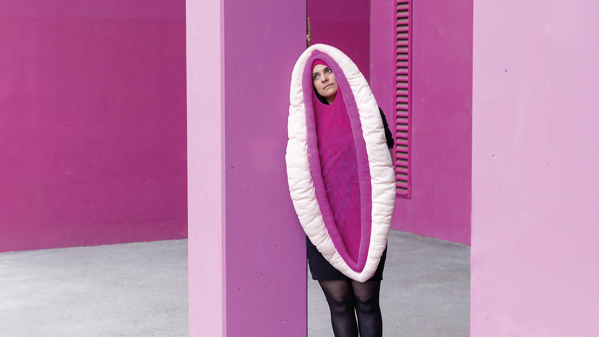 Vulva: Performancekostüm komplett sichtbar – Eine Frau lehnt an einer pinken Wand, sie trägt ein großes, pink-weißes Kostüm in ovaler Form, welches aussieht wie eine Vulva.