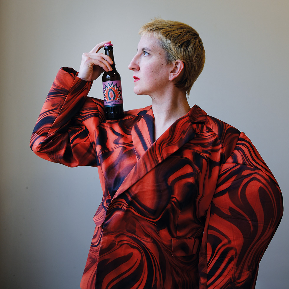 Vulva: Person mit Bierflasche – Eine Frau in rotem gemusterten Outfit hält eine Flasche „Muschicraft“-Bier an die Schulter.