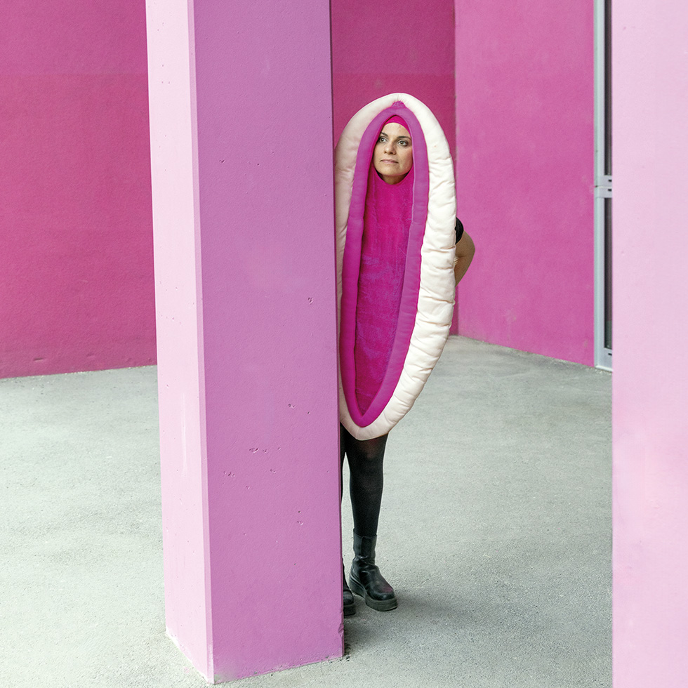 Vulva: Performancekostüm rosa – Eine Frau steht hinter einer pinken Säule, sie trägt ein großes, ovales Kostüm in Rosa und Pink, dass aussieht wie eine Vulva