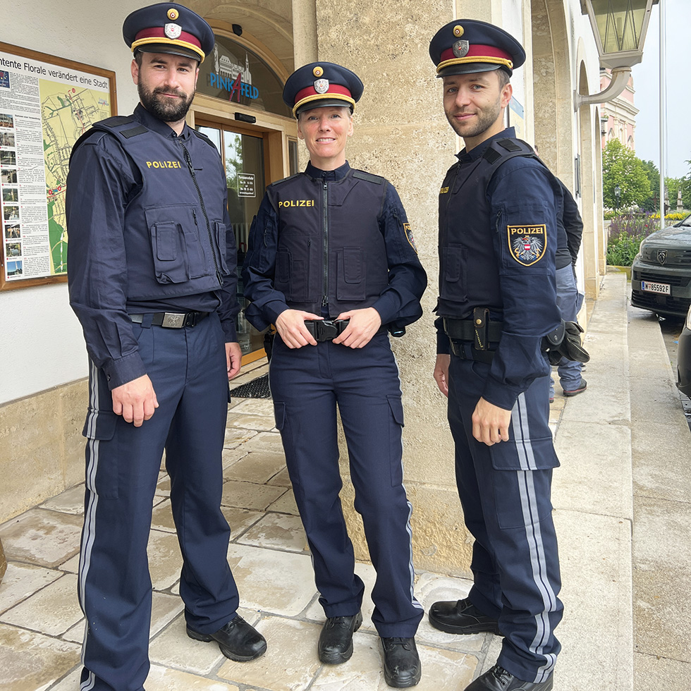 Drei Polizist:innen – Zwei Männer und eine Frau in dunkelblauen Polizeiuniformen stehen vor einem Gebäudeeingang und blicken in die Kamera.
