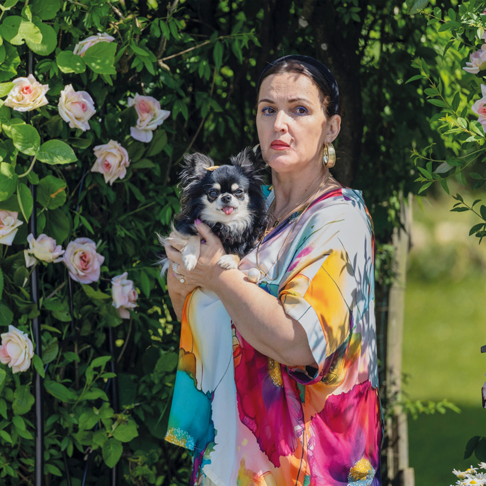 Zuagroast: Frau mit Hund – Eine Frau in bunter Bluse hält einen kleinen schwarz-weißen Hund vor Rosenbüschen.