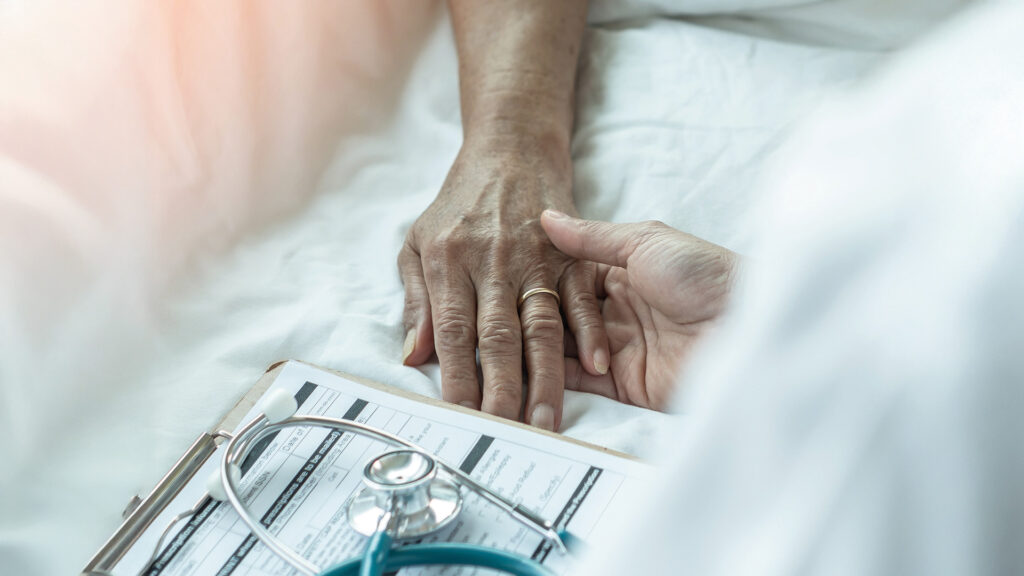Assistierter Suizid: Arzt oder Pfleger hält fürsorglich die Hand einer älteren Person im Krankenhausbett