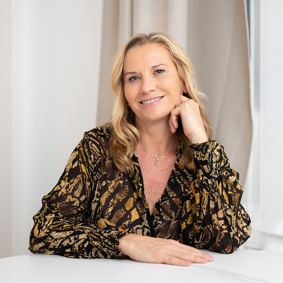 Lächelnde Frau mit blondem Haar in gemusterter Bluse sitzt an einem Tisch und blickt in die Kamera.