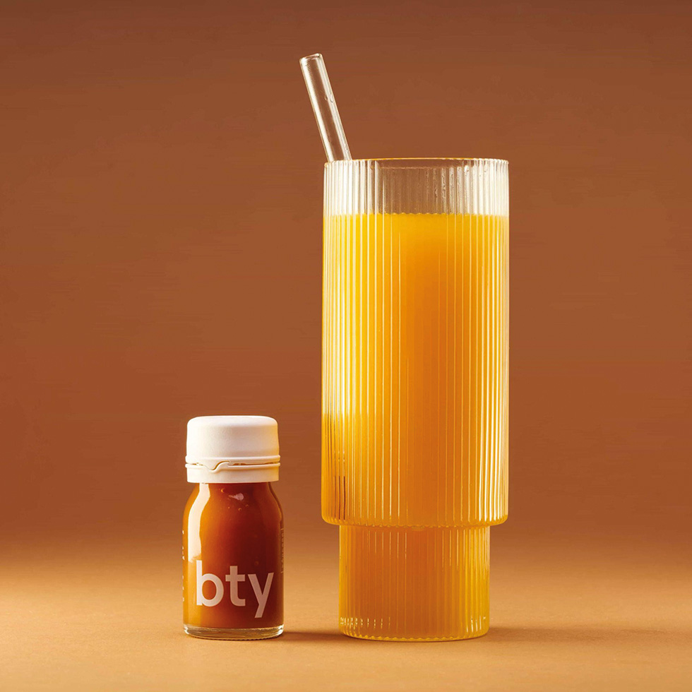 Glas mit Glastrinkhalm und oranger Flüssigkeit, daneben eine kleine Schraubflasche mit dem Aufdruck "bty" auf braunem Hintergrund