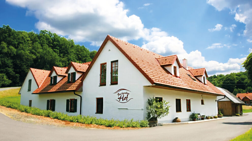 Dorothea Jagschitz: Traditioneller Bauernhof mit rotem Dach und weißer Fassade in idyllischer Landschaft