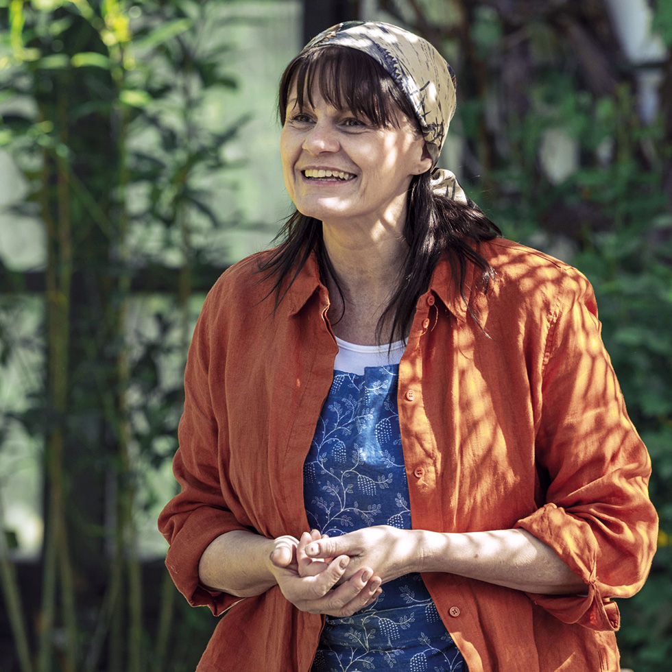 Frau im Garten – Eine Frau mit Kopftuch und orangefarbener Bluse lächelt, Pflanzen im Hintergrund.