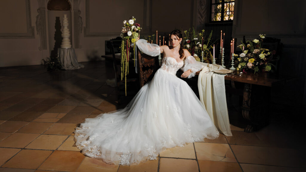 Braut in romantischem Spitzenkleid mit ausladender Schleppe, sitzend an festlich gedeckter Hochzeitstafel