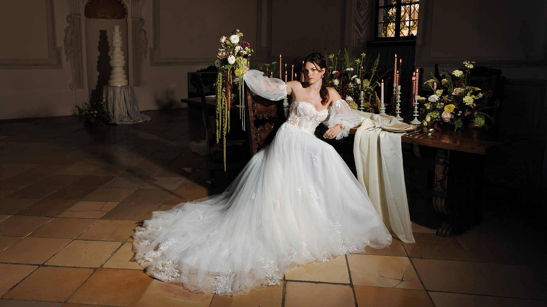 Braut in romantischem Spitzenkleid mit ausladender Schleppe, sitzend an festlich gedeckter Hochzeitstafel