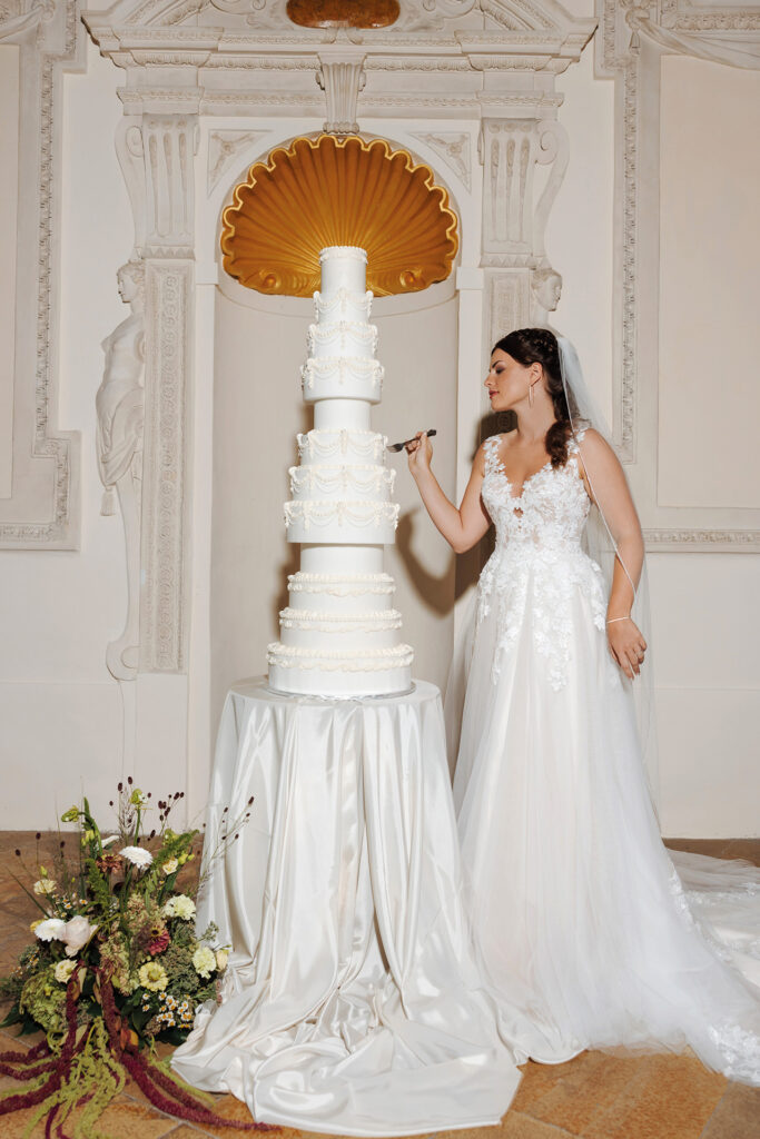 Braut im Spitzenkleid neben imposanter, mehrstöckiger Hochzeitstorte – stilvolles Hochzeitsshooting
