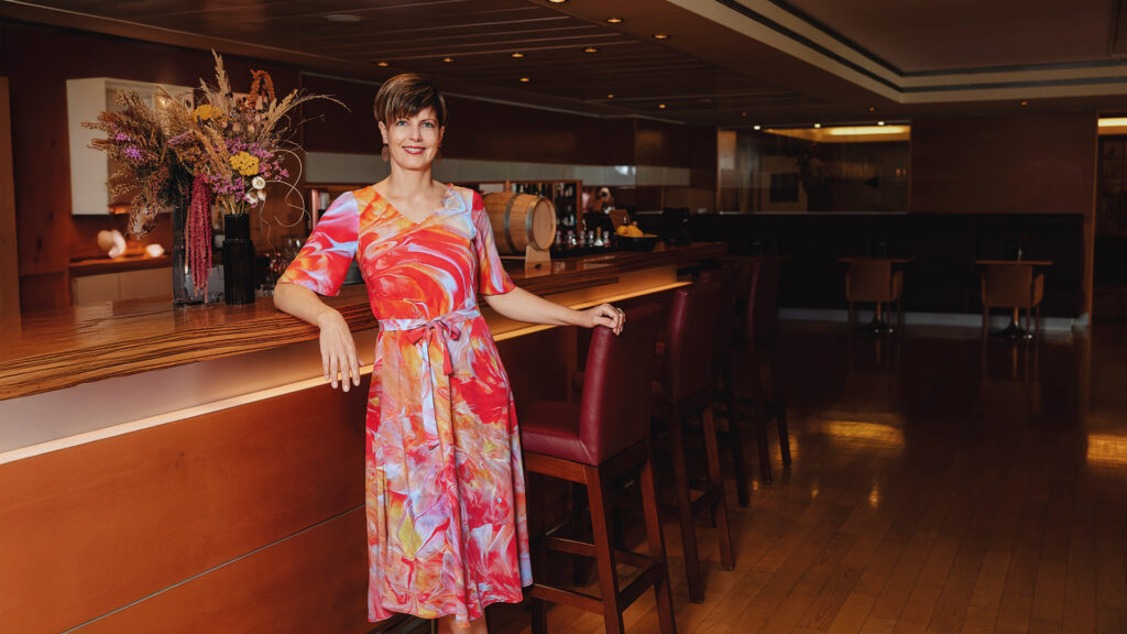 Kunst: Kurzhaarige Frau in einem bunt gemusterten Jersey-Kleid leht an einer Holzbar und lächelt in die Kamera