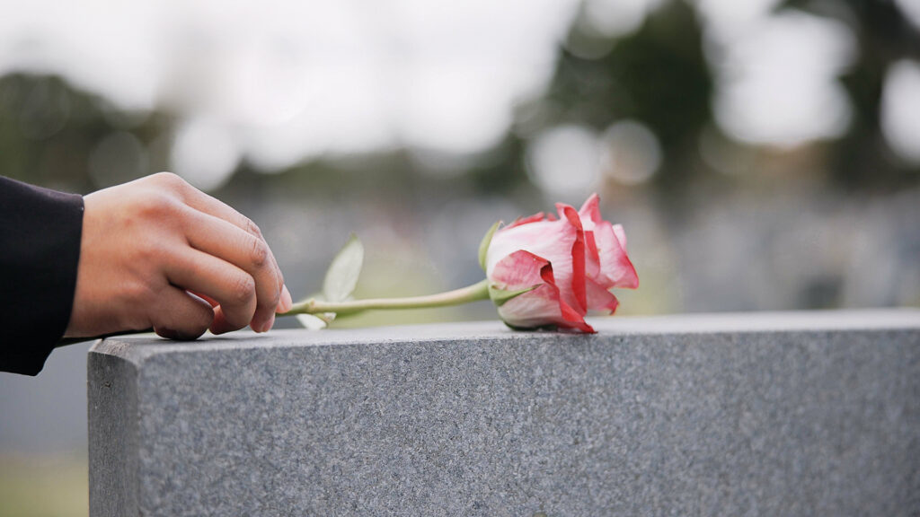 Hand legt eine einzelne Rose auf einen Grabstein, Symbol des Abschieds.