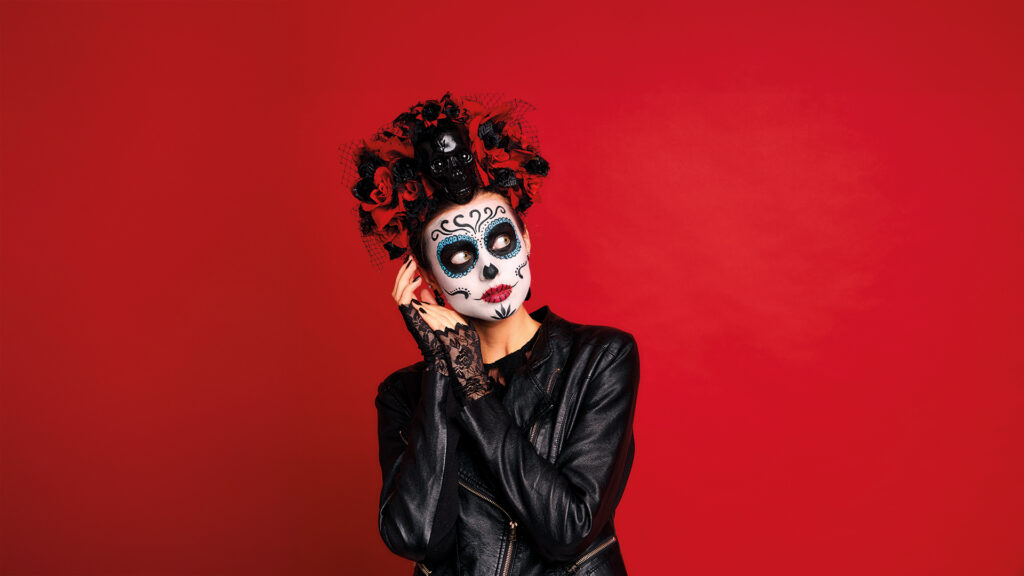 Abschied: Person mit kunstvollem Totenkopf-Make-up und Kopfschmuck aus roten Rosen vor rotem Hintergrund.