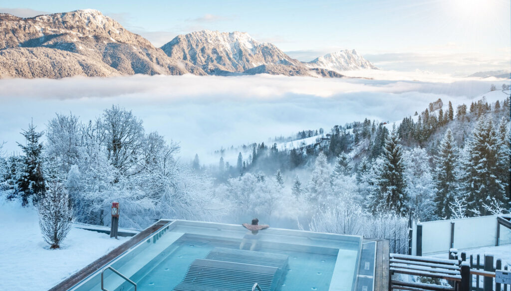 Panorama Outdoor-Pool in den Bergen bei einer verschneiten Winterlandschaft mit einem Wolkenmeer im Tal im Natur- & Wellnesshotel Höflehner ****Superior in der Region Schladming-Dachstein.
