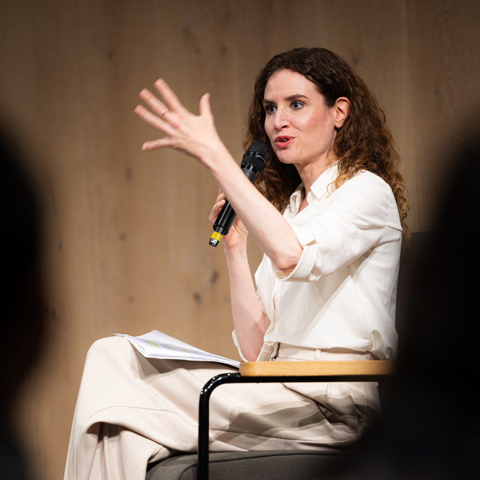 Judith Kohlenberger: Frau in weißem Outfit spricht gestikulierend in ein Mikrofon.