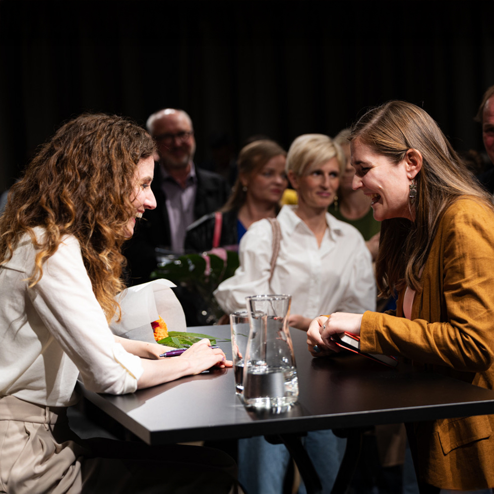 Judith Kohlenberger: Frau signiert ein Buch und unterhält sich lächelnd mit einer Besucherin, im Hintergrund weitere Gäste.