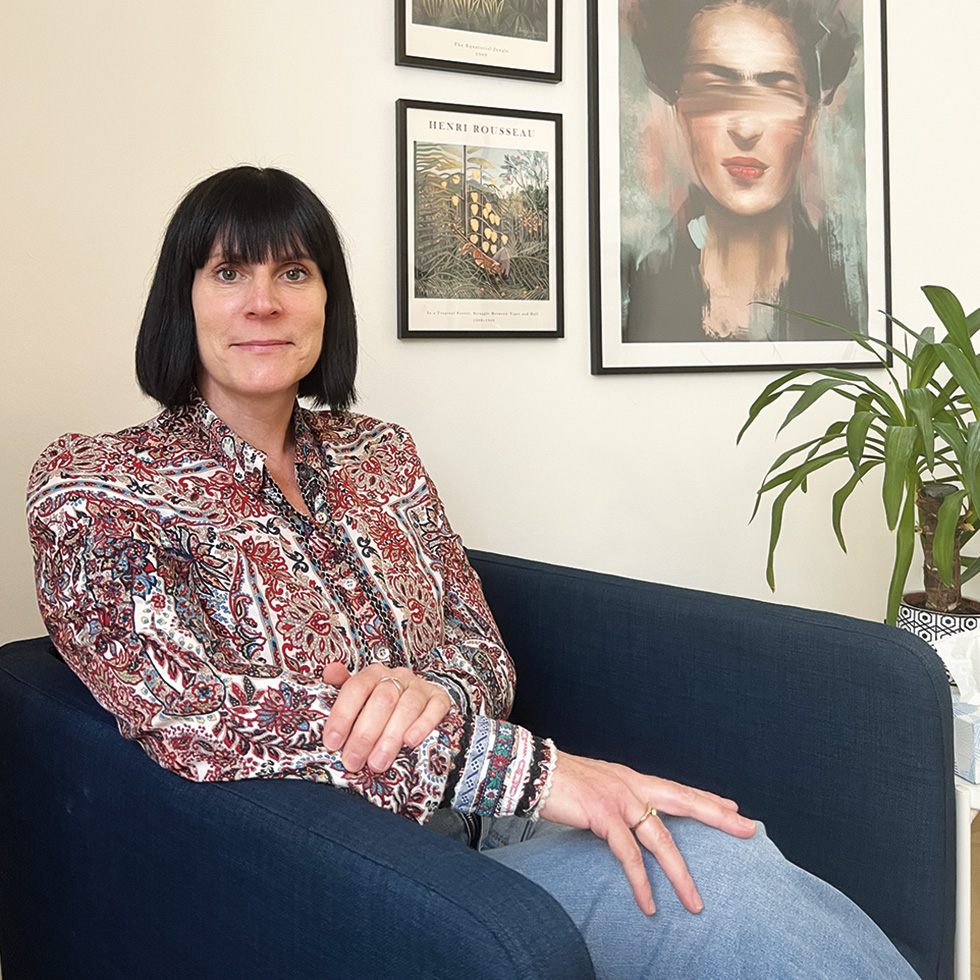 Cybergewalt: Frau mit dunklem Haar und gemusterter Bluse sitzt in einem Sessel, im Hintergrund Kunstwerke und Pflanzen.